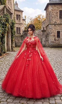 A woman stands on a cobblestone path before stone buildings, wearing the Quinceanera Dress CHF3012 By Chicas in vibrant red, featuring 3D floral accents and a dramatic ballgown skirt. Her hair is elegantly styled in an updo, and a matching cape is gracefully draped over her shoulders.