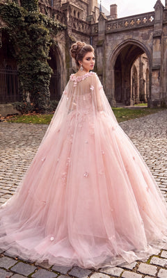 Wearing the Quinceanera Dress CHF3012 By Chicas, a woman stands on a cobblestone path in front of a stone castle or cathedral. The dress features a pink ballgown skirt and is paired with a sheer cape adorned with 3D floral accents.