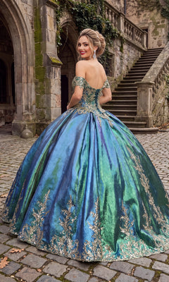 A woman wearing the Quinceanera Dress CHF3015 By Chicas—a blue and green off-shoulder ball gown adorned with gold embellishments—stands on a stone path before an old stone building featuring stairs and arches.