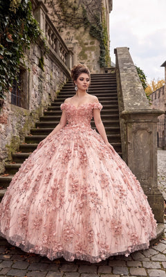 A woman in the Quinceanera Dress CHF3017 by Chicas, a blush off-the-shoulder ball gown with 3D floral appliques, stands on a stone staircase before an ivy-covered historic building, embodying the timeless elegance of a classic quinceanera ensemble.