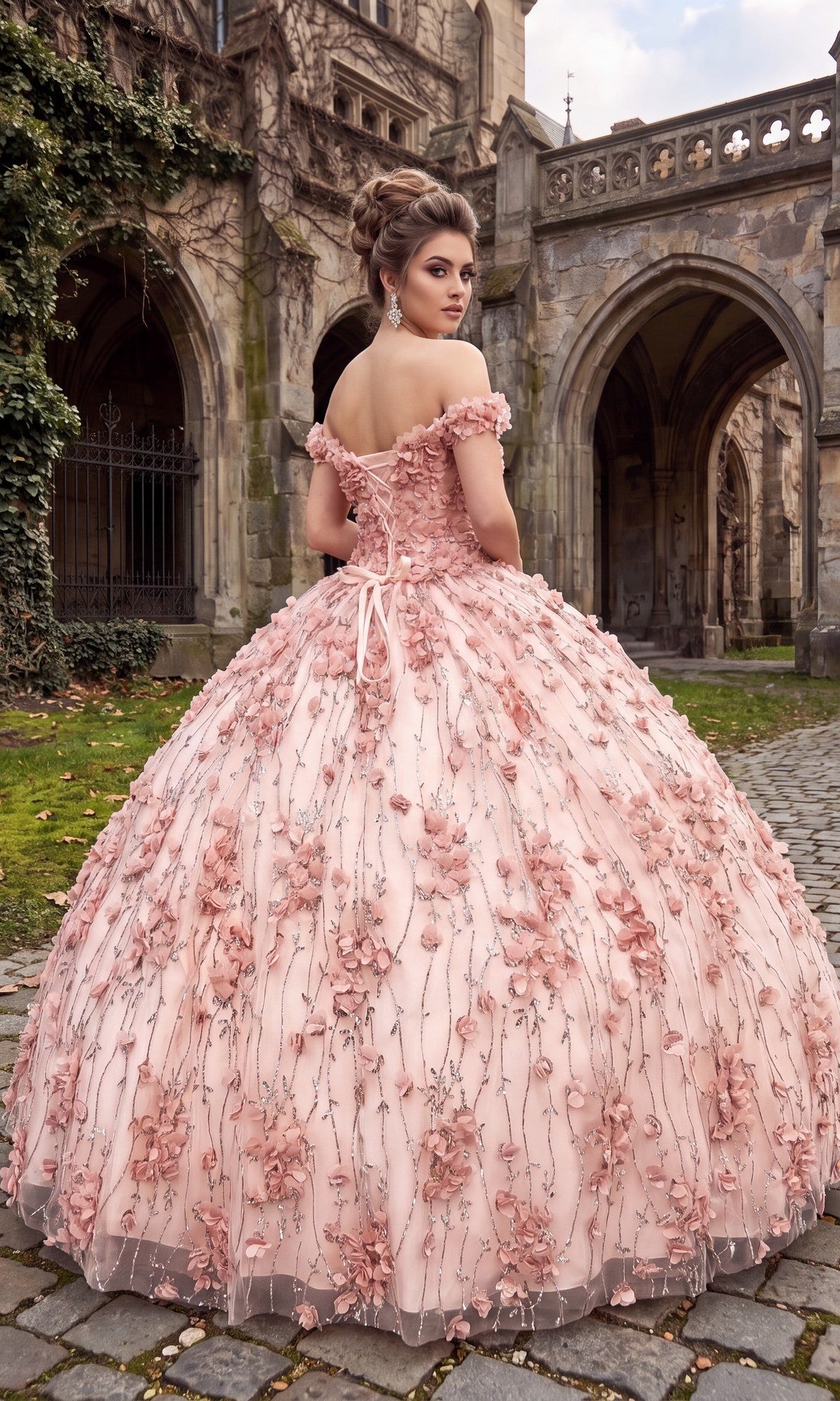 A woman wearing the Quinceanera Dress CHF3017 By Chicas—an elaborate, off-the-shoulder gown adorned with 3D floral appliques—stands on cobblestones in front of a historic stone building.