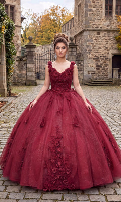 A woman in the Quinceanera Dress CHF3019 by Chicas, a burgundy glitter-tulle ballgown, stands on a cobblestone path outside a stone building with autumn trees in the background.