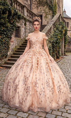 Wearing the Quinceanera Dress CHF3020 By Chicas in blush, a woman stands on a cobblestone path beside ivy-covered stone steps and an old building; her gown features an off-the-shoulder neckline with delicate 3D flower appliqué.