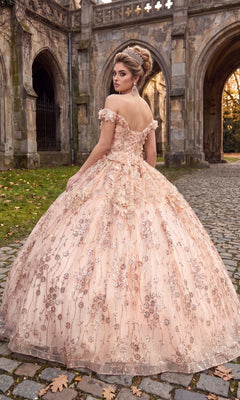 Wearing the Quinceanera Dress CHF3020 By Chicas, a woman stands on cobblestone near a stone building, looking over her shoulder in an ornate blush gown with 3D flower appliqué and an off-the-shoulder neckline.