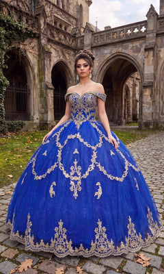 A woman in the Quinceanera Dress CHF3025 by Chicas, featuring royal and gold hues, stands on a cobblestone path in front of a historic stone building.