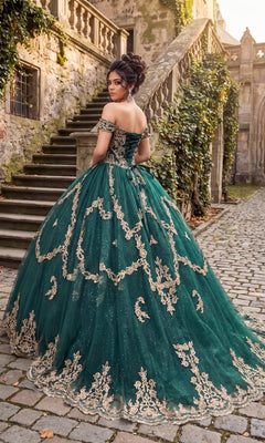 A woman wearing the Quinceanera Dress CHF3025 By Chicas, an emerald green off-the-shoulder gown featuring intricate gold embroidery, stands on a cobblestone path near stone stairs and ivy-covered walls, gazing over her shoulder.