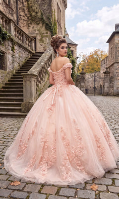 Wearing the Quinceanera Dress CHF3034 By Chicas—an elegant off-the-shoulder ballgown adorned with delicate floral appliqués—a woman stands on a cobblestone path near stone stairs and historic buildings, her back to the camera as she gracefully turns her head over her shoulder.