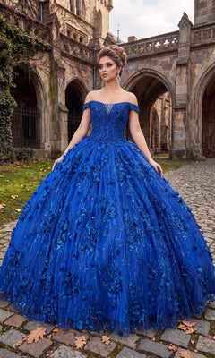 A woman wearing the Quinceanera Dress: Chicas CHF3038 in Royal stands on a cobblestone path in front of a historic stone building, the 3D floral appliques and tulle ballgown skirt cascading gracefully around her.