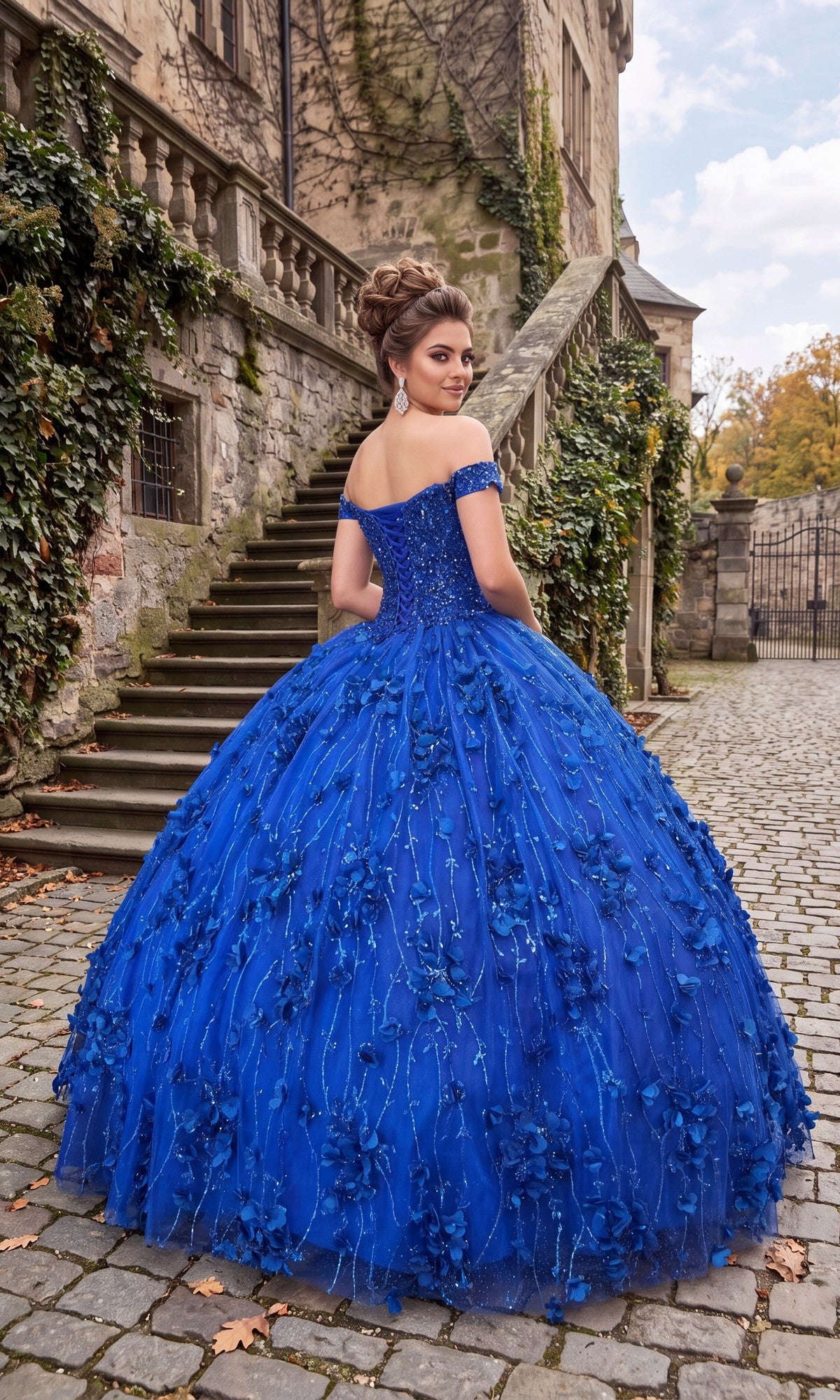 Wearing the Chicas CHF3038 Quinceanera Dress, a woman stands on cobblestone near stone steps and an ivy-covered building. The bright blue gown features a voluminous tulle ballgown skirt and intricate 3D floral appliques as she looks back over her shoulder.