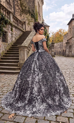 Wearing the Quinceanera Dress CHF3039 By Chicas, a woman stands on a cobblestone path near stone stairs and historic buildings, looking over her shoulder. The elegant black ballgown features intricate silver embroidery for a timeless and sophisticated look.