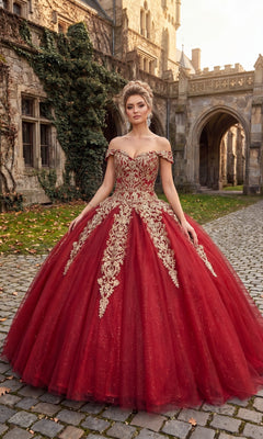 A woman stands outdoors on cobblestone in front of a stone building, wearing the Quinceanera Dress CHF3040 By Chicas in burgundy and gold, featuring an off-the-shoulder design with gold embroidery and a glitter tulle ballgown skirt.