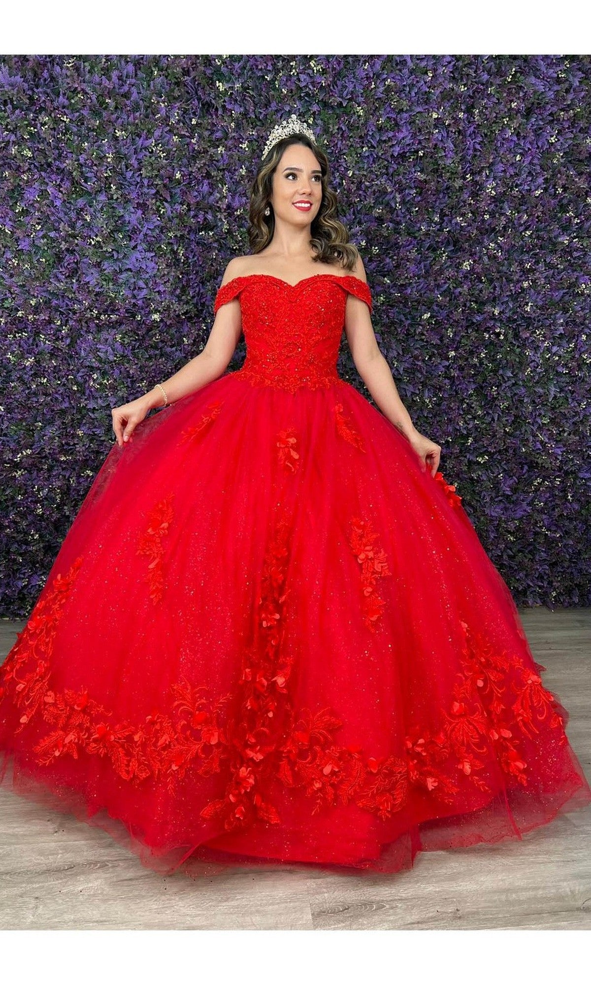 A woman wearing the Quinceanera Dress CHF3041 By Chicas in red, featuring off-the-shoulder floral details, stands smiling before a purple flower wall and holds the sides of her ballgown skirt.