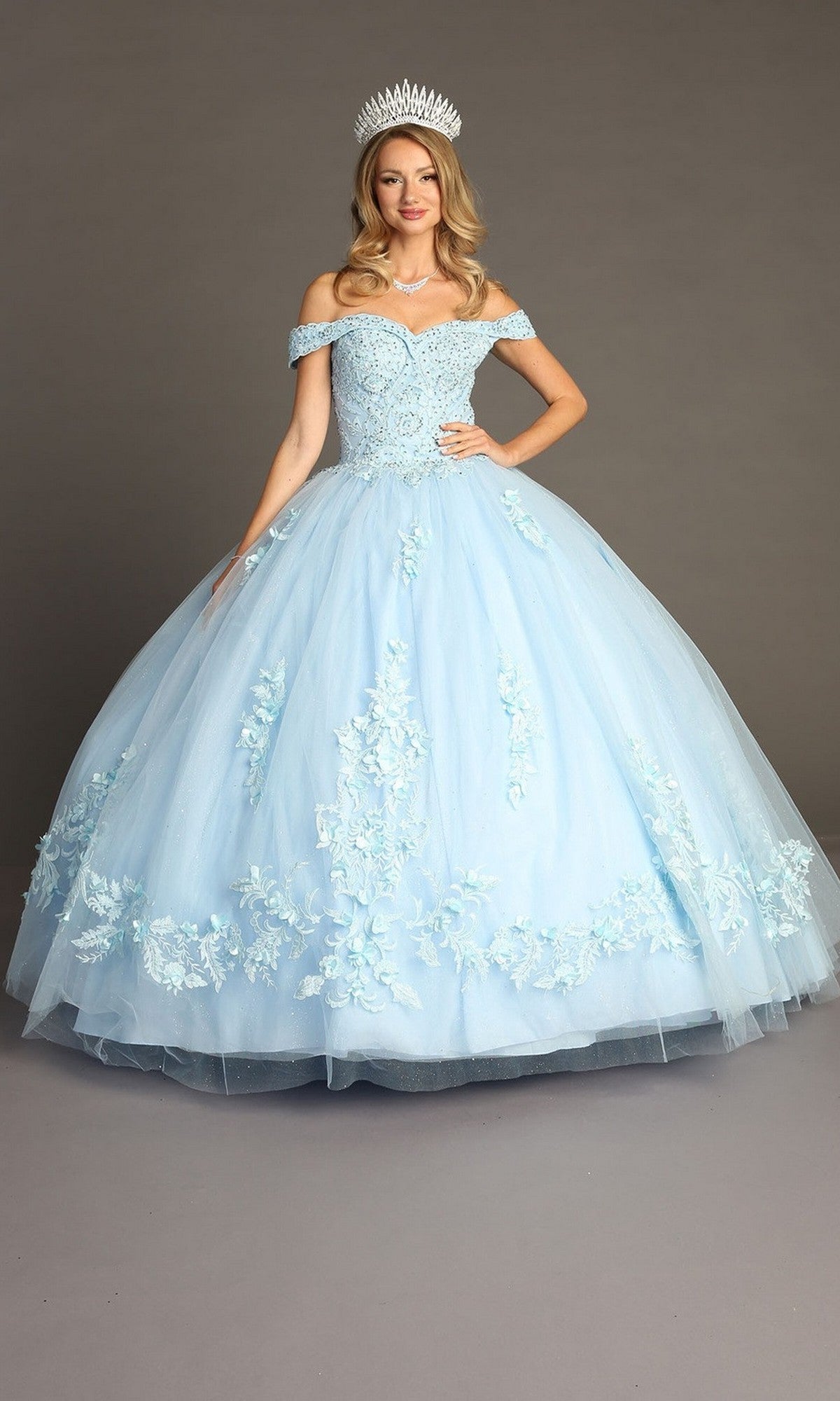 A woman stands against a plain background wearing the Quinceanera Dress CHF3041 by Chicas in sky blue—an off-the-shoulder ballgown skirt adorned with delicate floral details and completed with a tiara.
