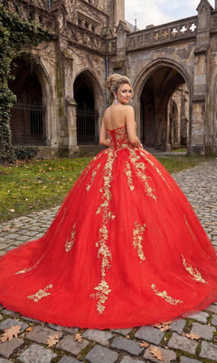 Wearing the breathtaking Quinceanera Dress CHF3042 By Chicas, a woman stands on a cobblestone path before a gothic-style stone building, looking back over her shoulder in the exquisite red and gold ballgown adorned with intricate gold embroidery.