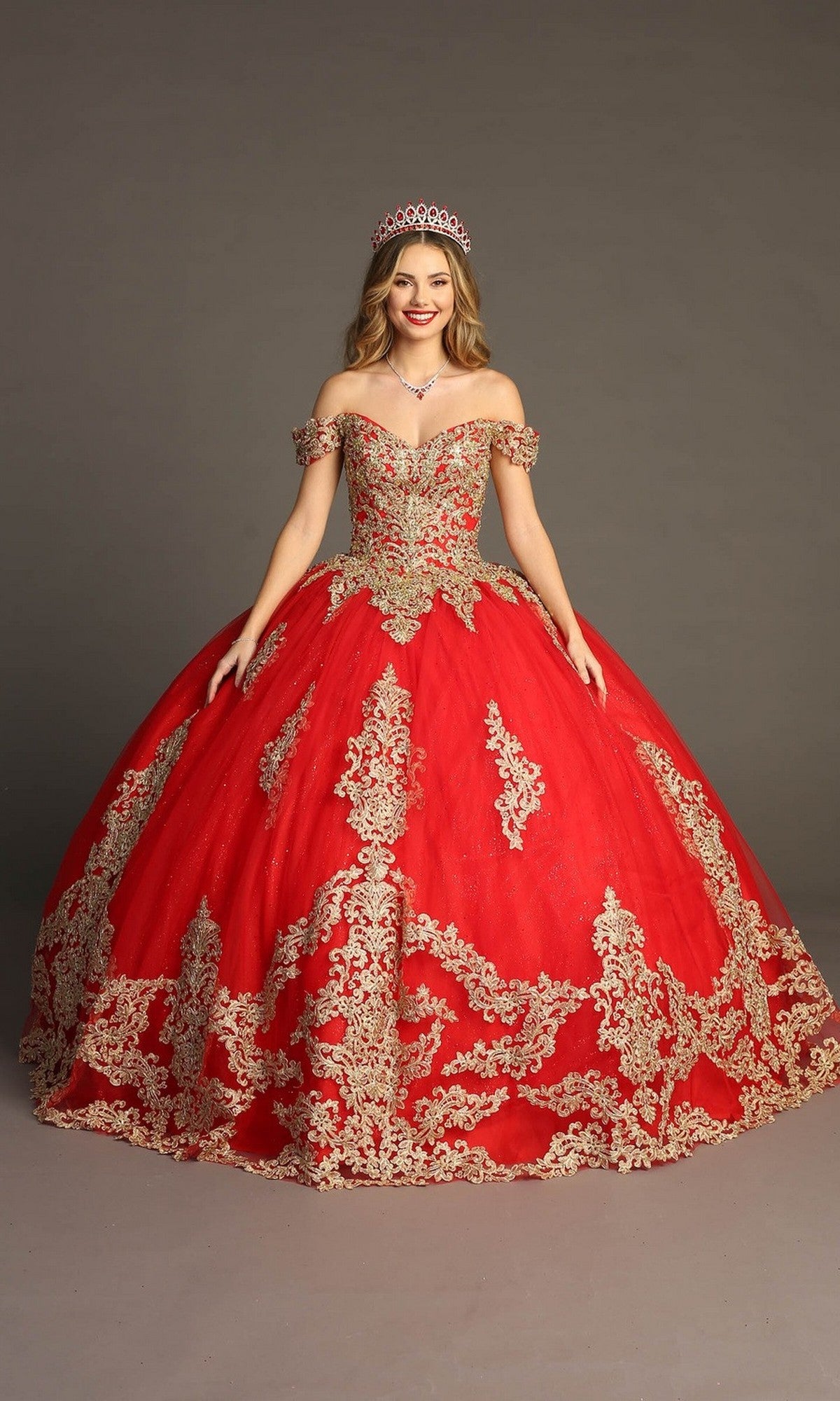 A woman wearing the Quinceanera Dress CHF3043 By Chicas in Red/Gold, with gold embroidery and a tiara, stands against a plain gray background, smiling at the camera.