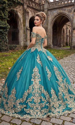 A woman in the Quinceanera Dress CHF3043 by Chicas, an ornate teal glitter tulle ballgown adorned with delicate gold embroidery, stands on a cobblestone path before an old stone building with arches.