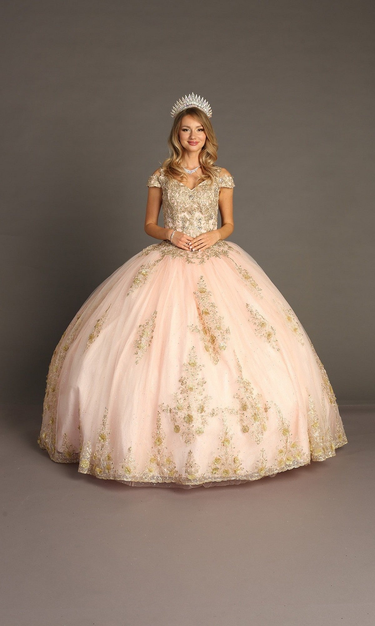 A young woman stands in a studio wearing the Quinceanera Dress CHF3051 by Chicas, an elegant pink gown adorned with gold embroidery and featuring a voluminous ballgown skirt; she clasps her hands gracefully in front of her as a sparkling tiara rests atop her head.