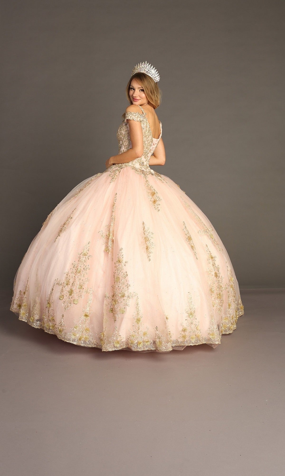Wearing the Quinceanera Dress CHF3051 By Chicas, a woman stands in front of a plain gray background, her back to the camera as she looks over her shoulder. The light pink off-the-shoulder gown features intricate gold embroidery and is complemented by a jeweled crown.