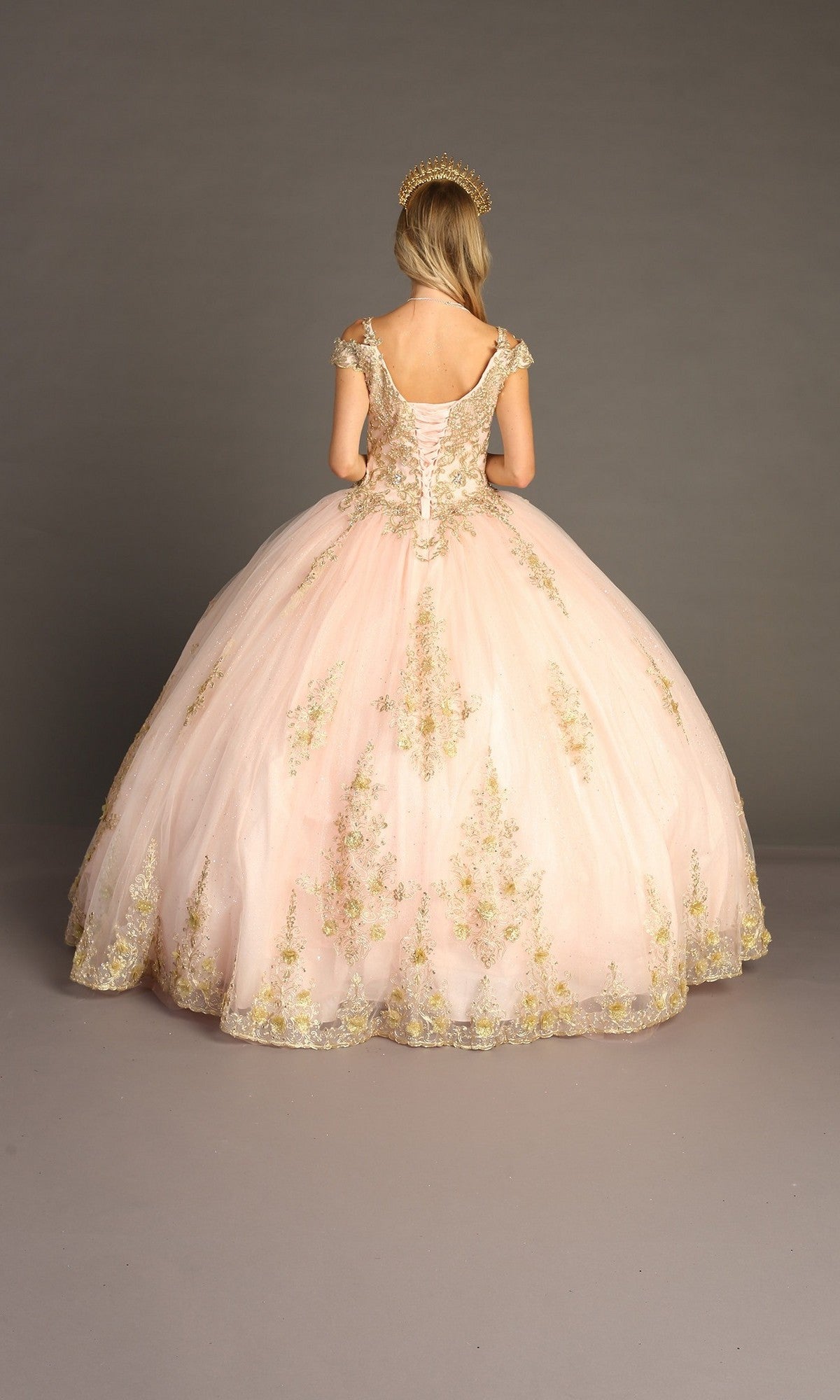 A person stands facing away against a plain background, wearing the Quinceanera Dress CHF3051 By Chicas—an off-the-shoulder pale pink ball gown adorned with gold embroidery and crowned with a tiara—evoking timeless Quinceanera elegance.