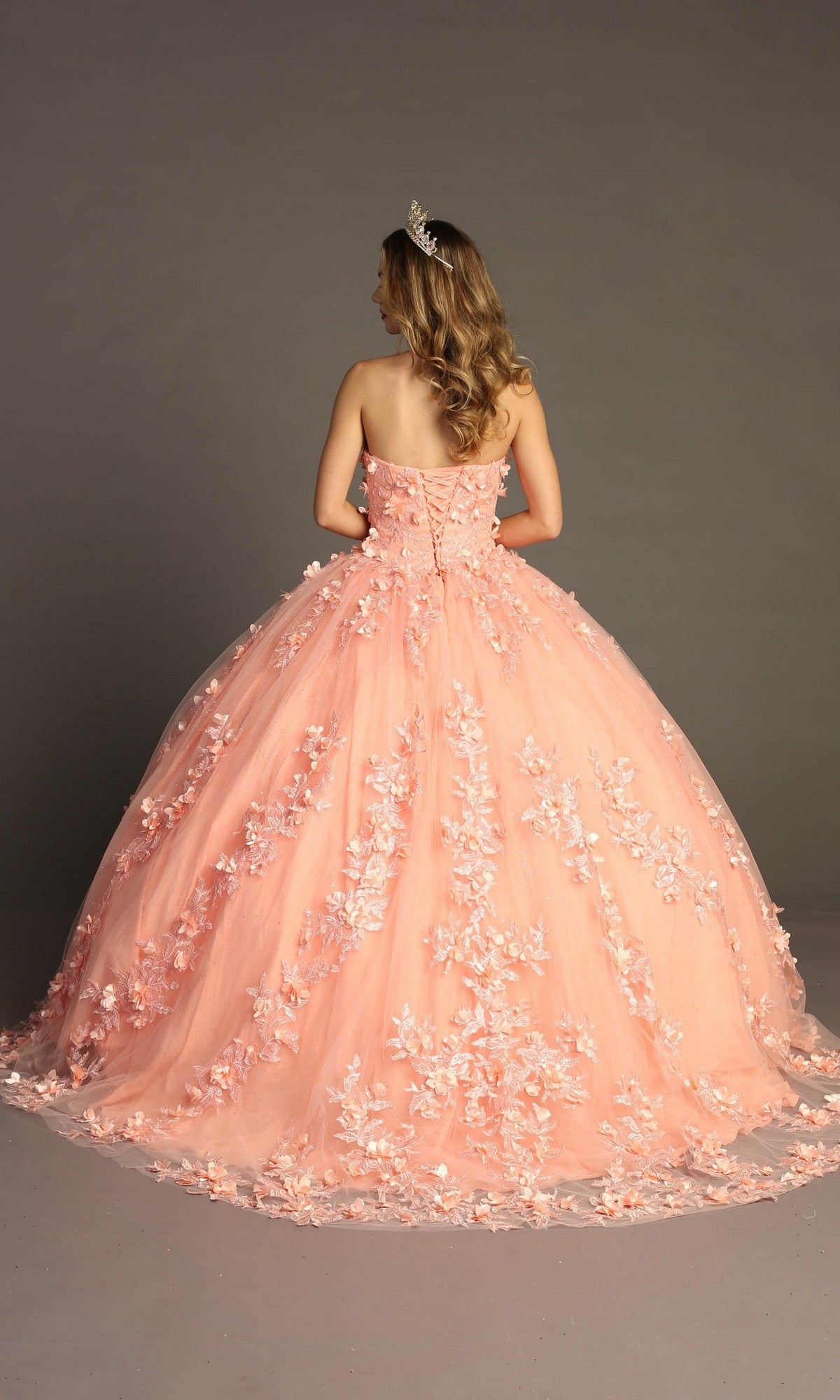 A woman wearing the Quinceanera Dress CHF3055 By Chicas—a peach strapless gown adorned with 3D floral appliqués—and a tiara stands facing away against a plain gray background.