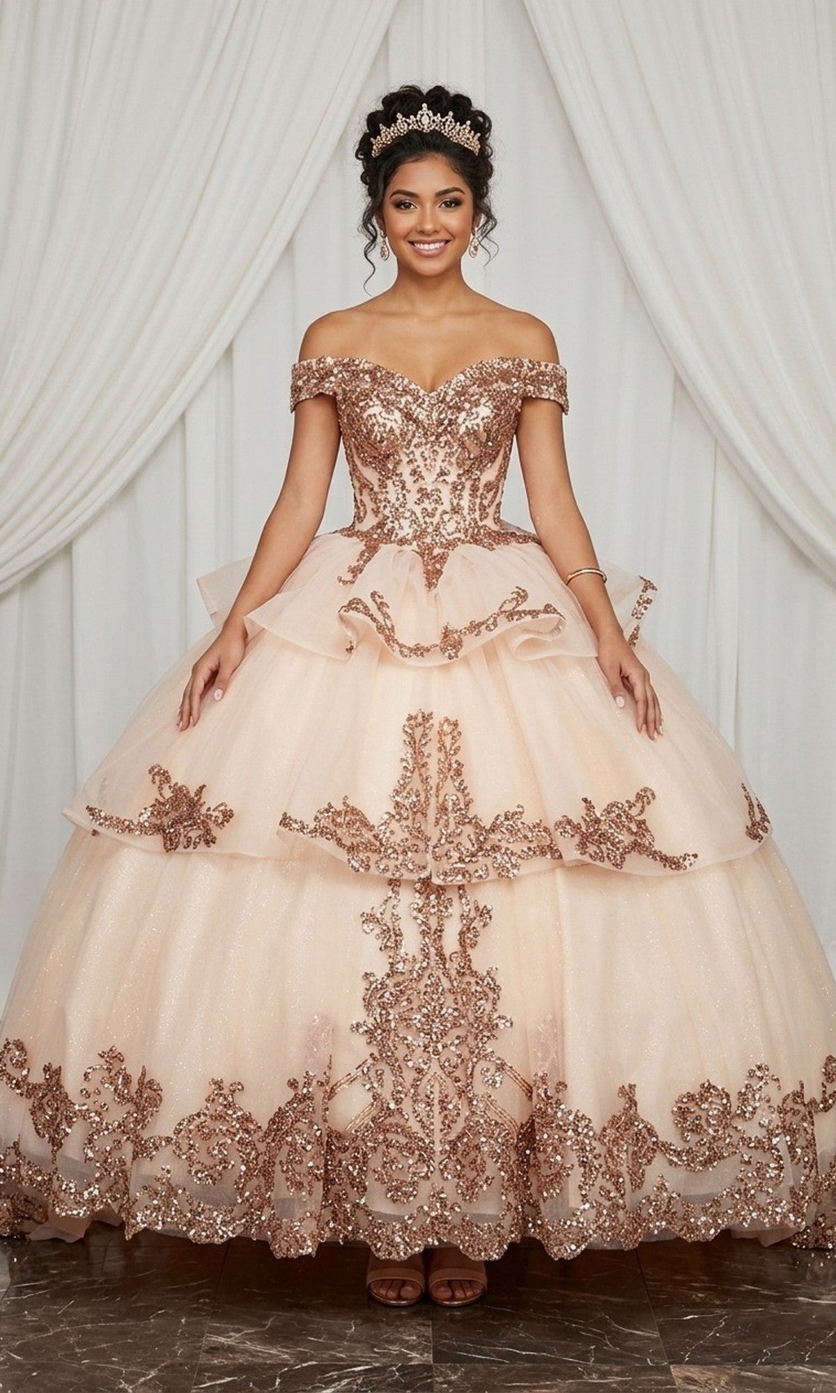 A young woman wearing the Quinceanera Dress CHF3063 by Chicas in champagne, featuring sparkly embellishments and an ornate off-the-shoulder design, stands in front of white drapes with a tiara atop her head.