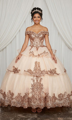 A young woman wearing the Quinceanera Dress CHF3063 by Chicas in champagne, featuring sparkly embellishments and an ornate off-the-shoulder design, stands in front of white drapes with a tiara atop her head.