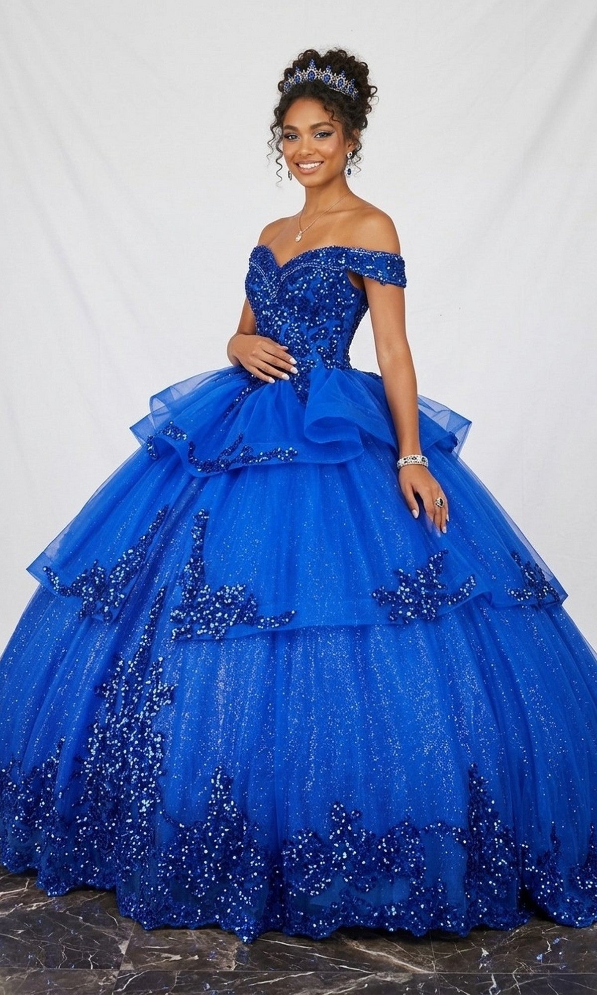 A woman stands against a white backdrop wearing the Quinceanera Dress CHF3063 By Chicas in royal, an off-the-shoulder gown with a full, sparkly tulle skirt and intricate beaded details. She completes her elegant look with a tiara and glamorous jewelry.