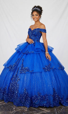 A woman stands against a white backdrop wearing the Quinceanera Dress CHF3063 By Chicas in royal, an off-the-shoulder gown with a full, sparkly tulle skirt and intricate beaded details. She completes her elegant look with a tiara and glamorous jewelry.
