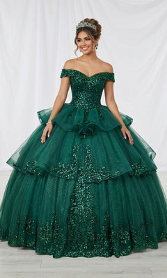 A woman wearing the Quinceanera Dress CHF3063 By Chicas in green, featuring a tiered ball gown silhouette with an off-shoulder neckline and sparkling sequin and tulle embellishments, stands against a white fabric backdrop.