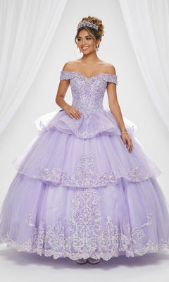 A young woman stands in front of white draped curtains, wearing the Quinceanera Dress CHF3063 By Chicas in lilac. The elegant off-the-shoulder gown features a tiered ball gown silhouette with a layered tulle skirt, intricate embroidery, and sparkly embellishments that catch the light. She completes her regal look with a delicate tiara.