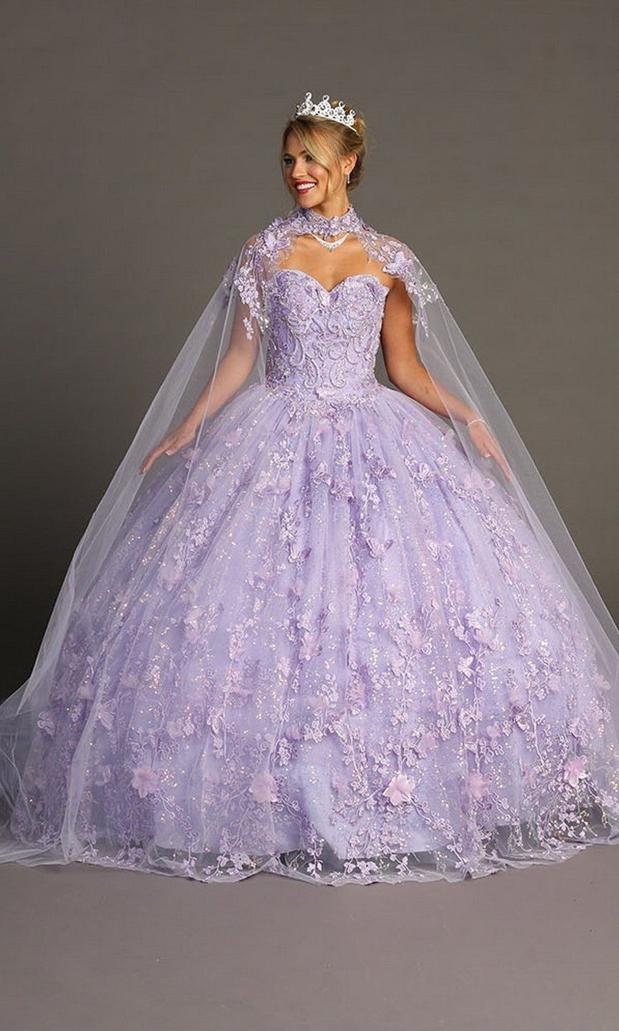 A woman wearing the Quinceanera Dress CHF3067 By Chicas in lilac, featuring a 3D floral-embroidered corset, a delicate sheer cape, and a tiara, stands against a plain gray background.
