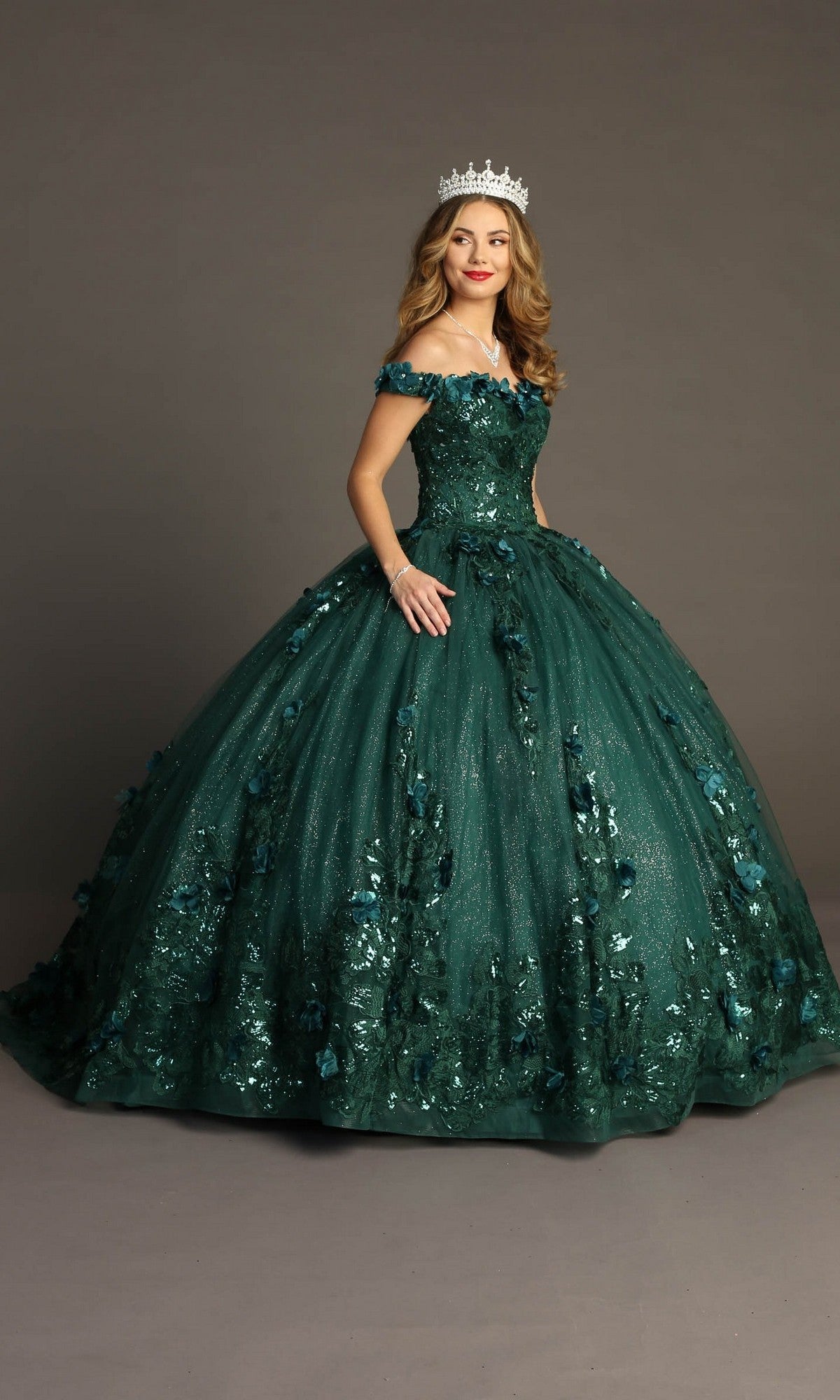A woman wearing a jeweled crown and the Quinceanera Dress CHF3069 By Chicas—an off-the-shoulder, dark green gown embellished with 3D floral appliques—stands against a plain background.