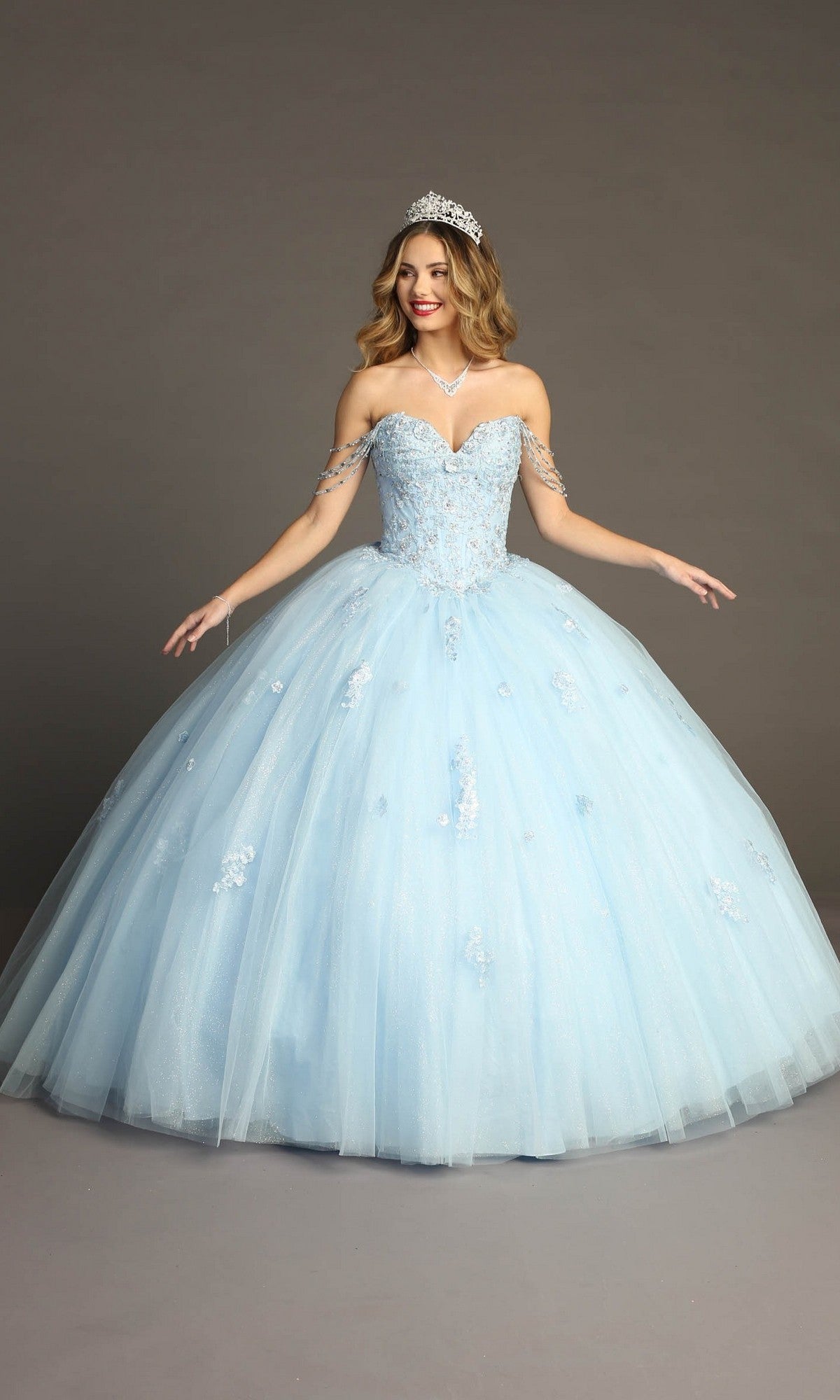 A woman stands against a plain gray background wearing the Quinceanera Dress CHF3071 By Chicas in baby blue—a glitter-tulle ballgown featuring off-shoulder straps, a beaded bodice for an elegant touch, a sparkling tiara, and a necklace.