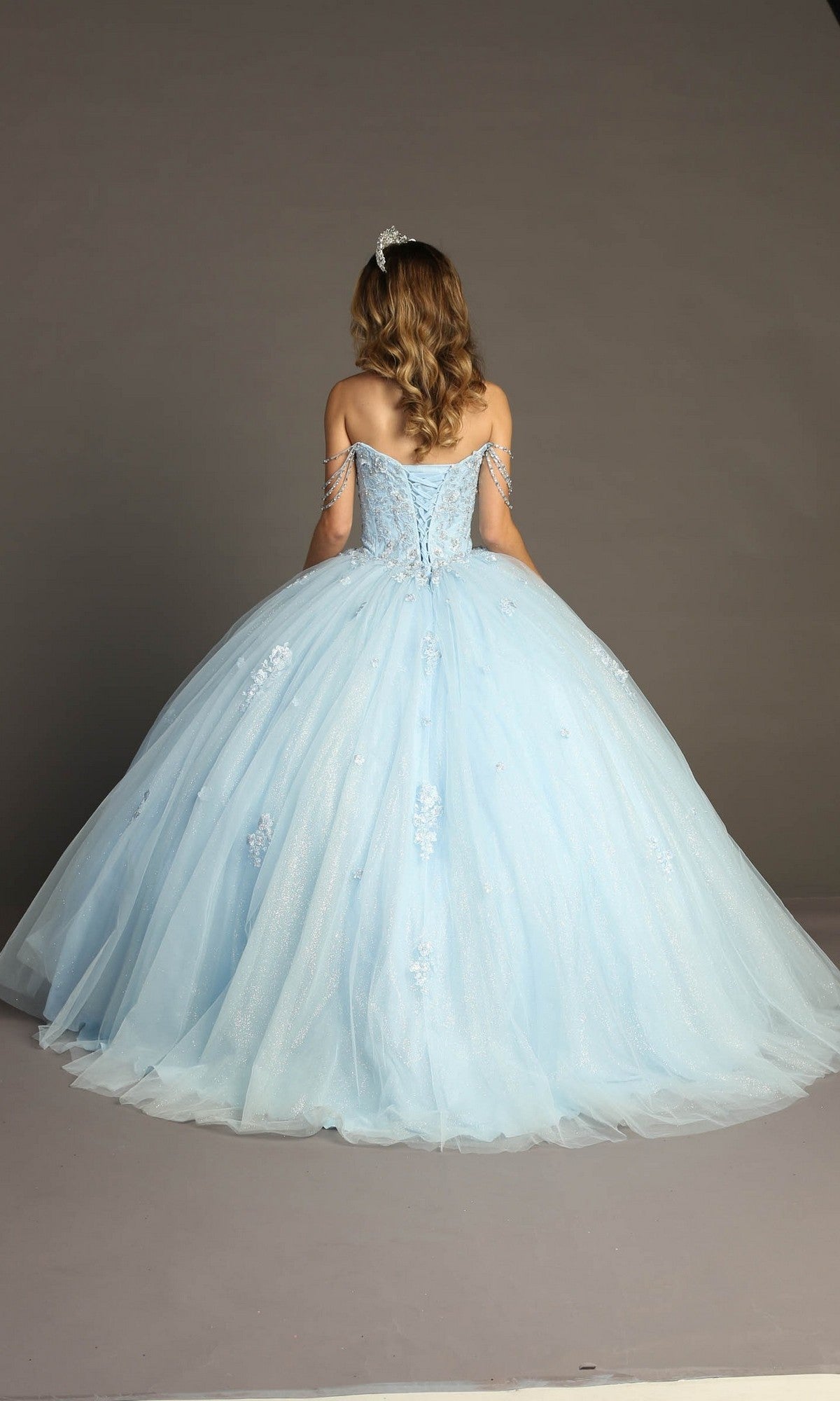 A woman with wavy hair stands facing away, wearing the Quinceanera Dress CHF3071 by Chicas—a light blue glitter-tulle ballgown with a full tulle skirt and off-the-shoulder straps—set against a plain gray background.