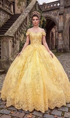 A woman wearing the Quinceanera Dress CHF3072 By Chicas in yellow, featuring intricate metallic embroidery and an ornate off-the-shoulder design, stands on a cobblestone path before a historic stone staircase and building, her dramatic ballgown skirt elegantly fanned around her.