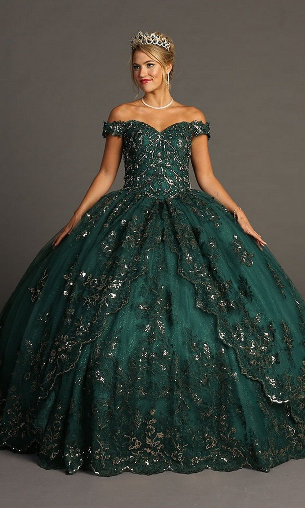 A woman stands against a plain gray background wearing the Quinceanera Dress CHF3072 By Chicas in green, an off-the-shoulder gown adorned with sequins, lace detailing, and exquisite metallic embroidery. She completes her look with a tiara, pearl necklace, and earrings.