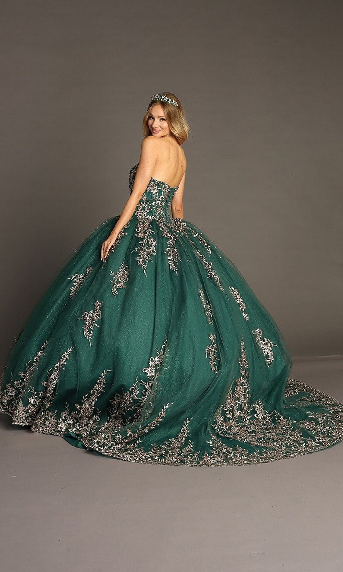 A woman is wearing the Quinceanera Dress CHF3073 By Chicas—a strapless dark green gown adorned with metallic-silver embroidery and silver embellishments, featuring a matching tiara and a flowing glitter-tulle skirt—as she stands against a plain backdrop.