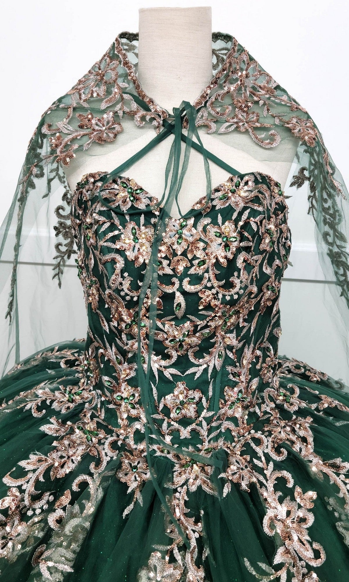 The Quinceanera Dress CHF3073 By Chicas is a strapless green ball gown adorned with gold and metallic-silver embroidery, complete with a matching sheer cape. Displayed on a dress form, this elegant gown features a glitter-tulle skirt that adds extra sparkle to its stunning design.