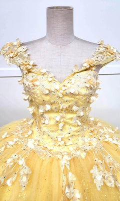 The Quinceanera Dress CHF3074 By Chicas is showcased on a mannequin, boasting a vibrant yellow off-shoulder design adorned with intricate lace embroidery. Its voluminous glitter tulle skirt is beautifully embellished with delicate beads and small fabric flowers.