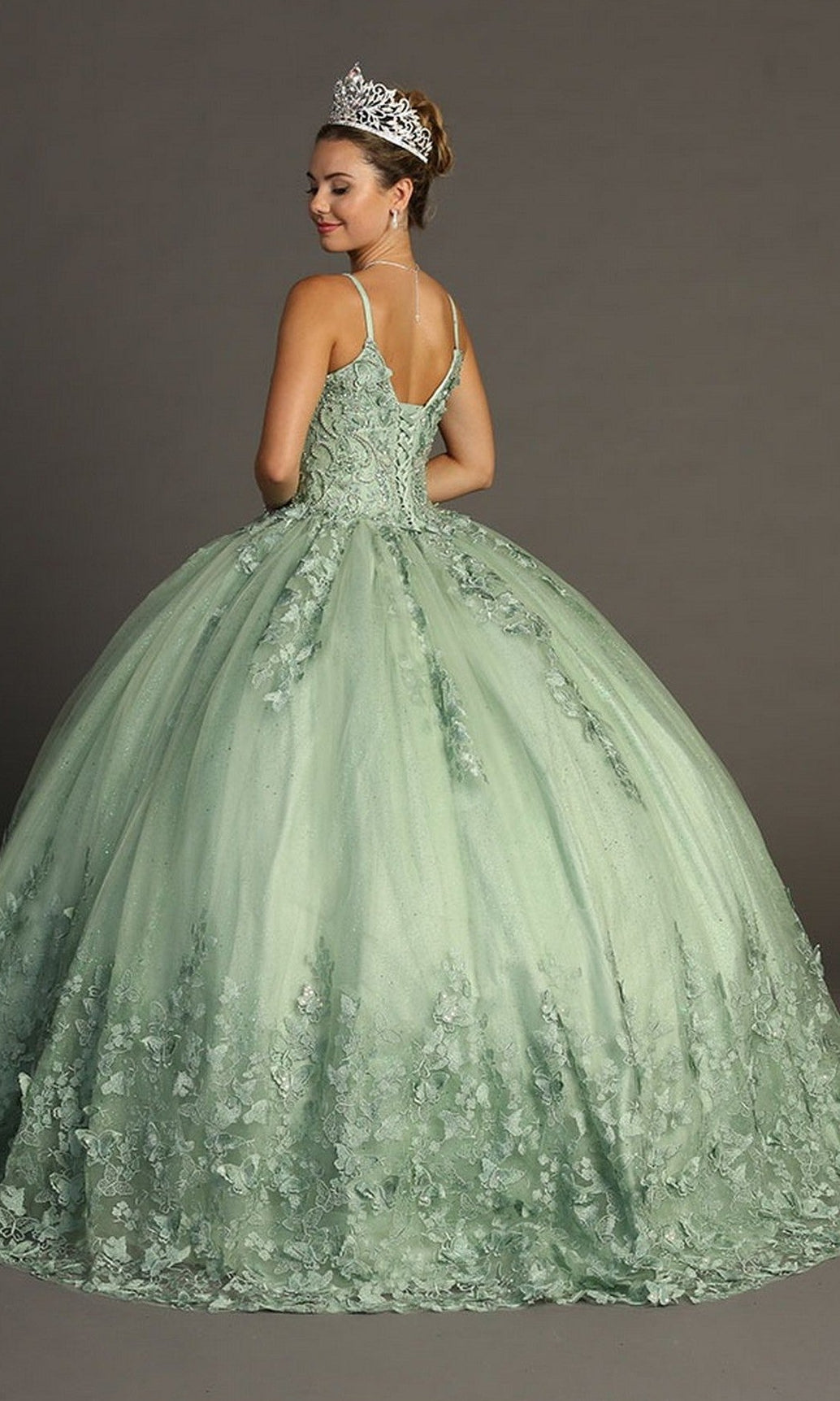 A woman wearing the Quinceanera Dress CHF3077 By Chicas—a light green glitter tulle ballgown adorned with delicate floral details and butterfly appliques—topped with a silver crown, stands against a plain gray background, facing away from the camera.