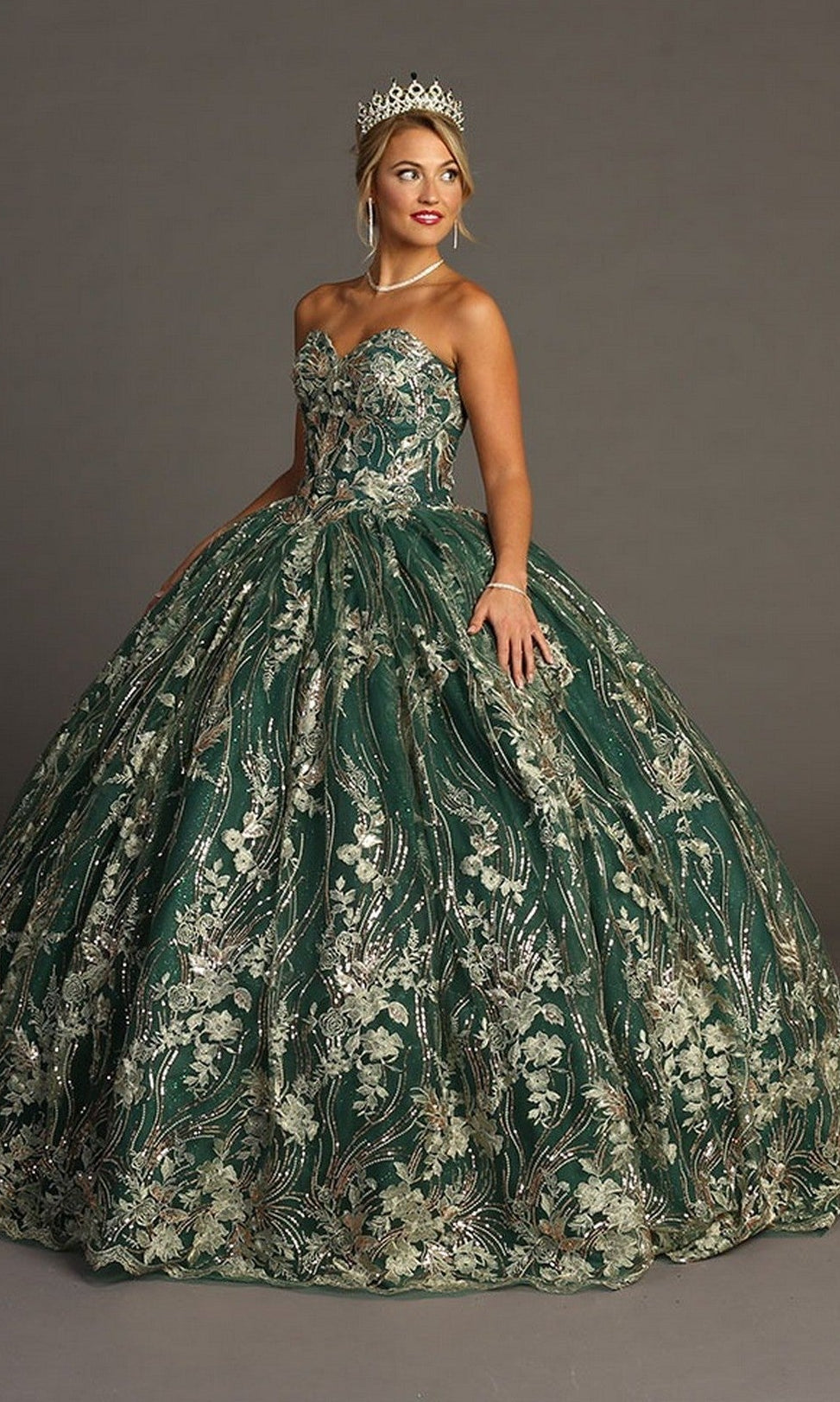 A woman stands against a plain gray background wearing the Quinceanera Dress CHF3079 By Chicas in Green/Gold—a strapless ballgown featuring metallic floral embroidery—accessorized with a tiara, necklace, and bracelet.