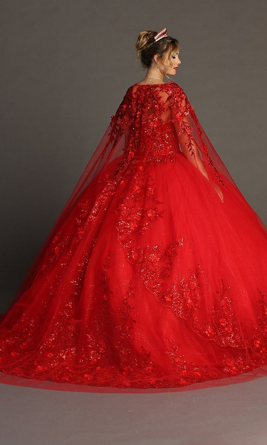 A woman stands facing away from the camera against a plain background, wearing the Quinceanera Dress CHF3080 By Chicas, featuring royal floral lace details and a sheer cape.