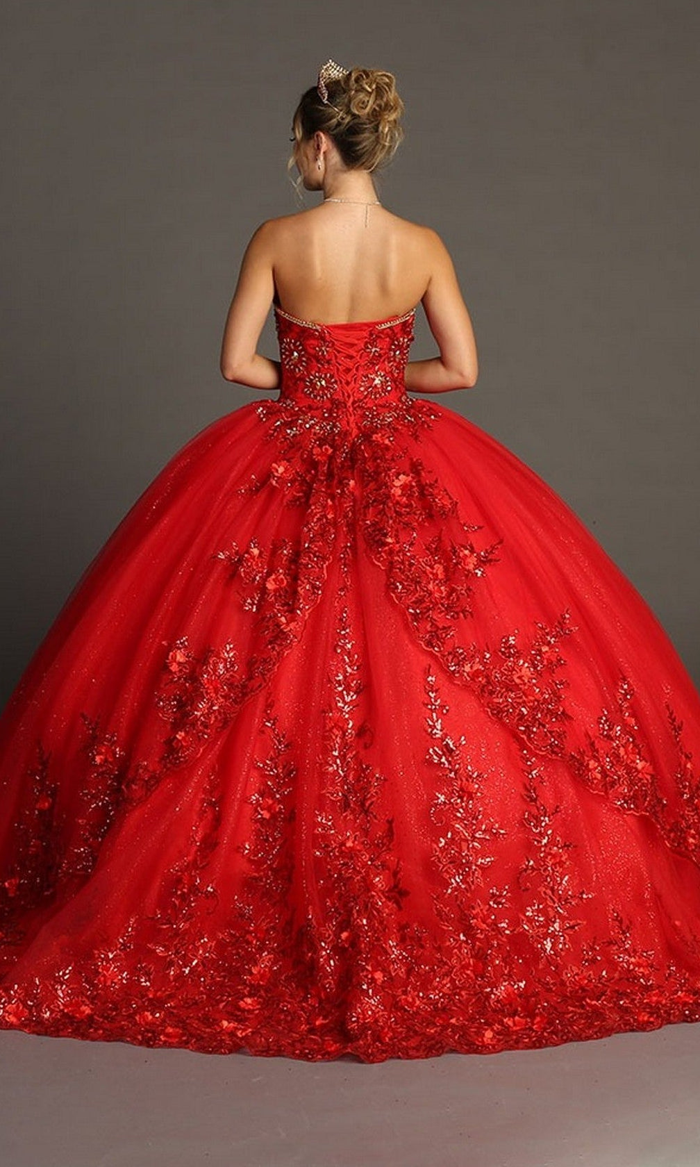 A woman stands facing away against a plain gray background, her hair in an updo adorned with a small tiara, wearing the Quinceanera Dress CHF3080 By Chicas—a strapless, floor-length red gown featuring 3D floral appliques.