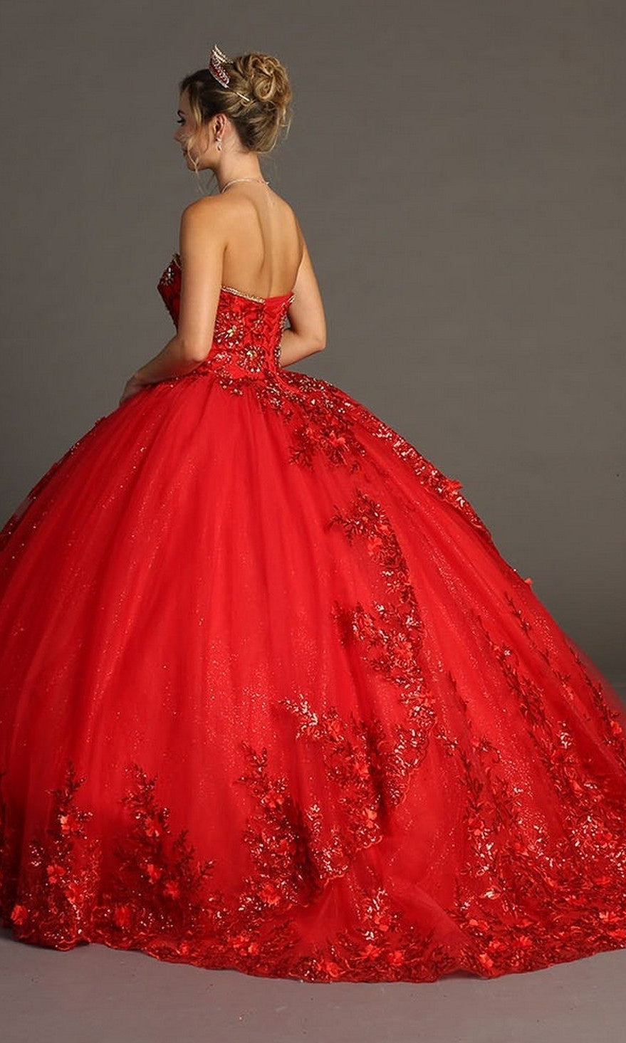 Woman wearing the Quinceanera Dress CHF3080 By Chicas, a strapless, bright red royal gown adorned with intricate 3D floral appliques, standing on a plain gray background, facing away from the camera.
