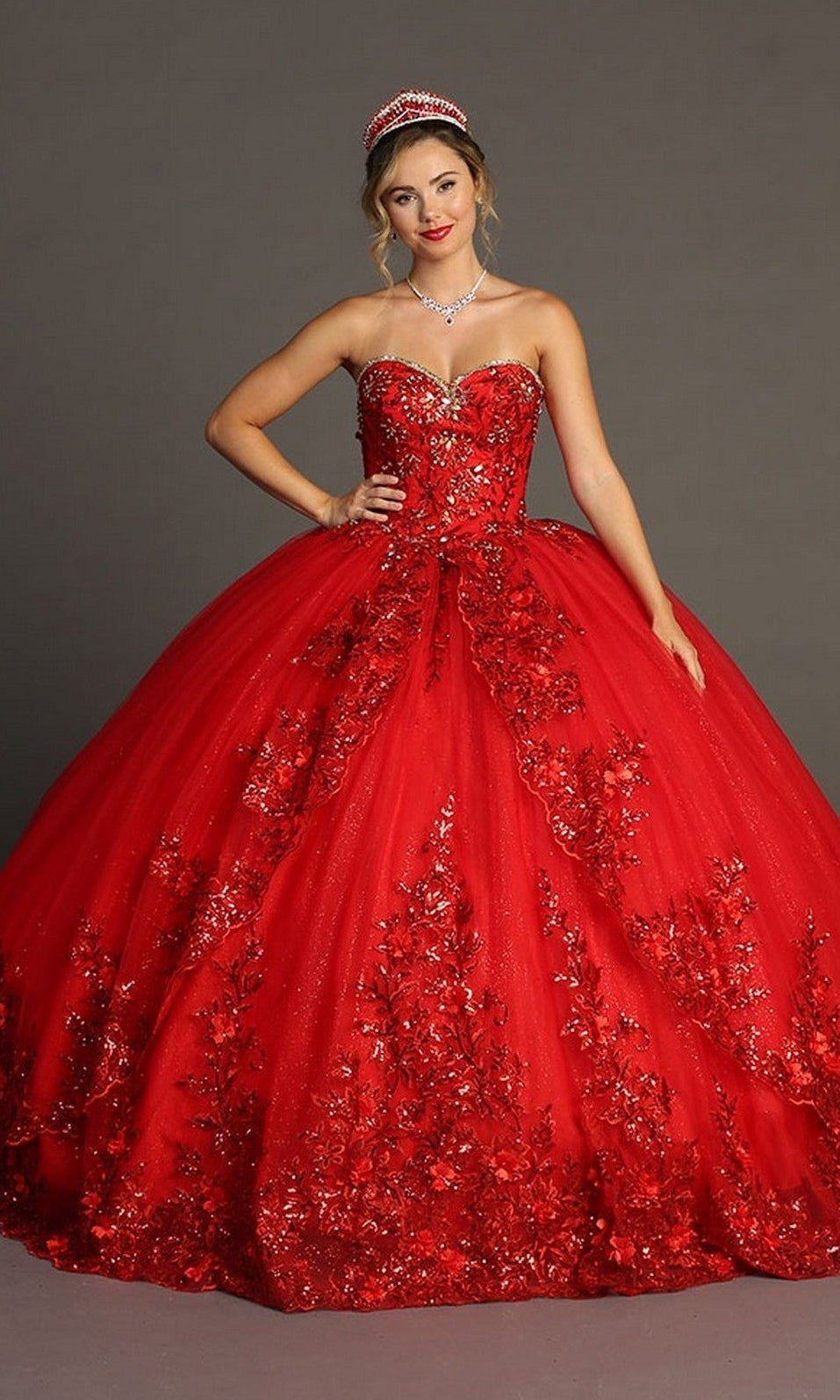 A woman wearing the Quinceanera Dress CHF3080 By Chicas, featuring red royal fabric embellished with 3D floral appliques, paired with a tiara and necklace, stands against a plain gray background.