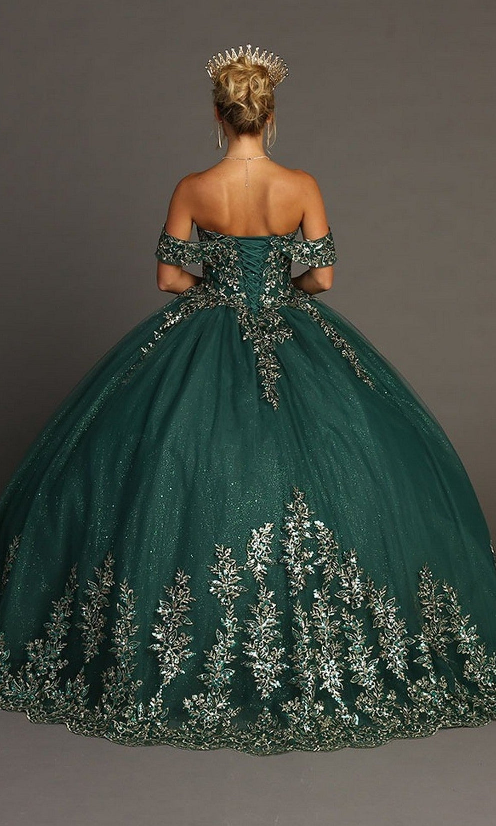 A woman is standing with her back to the camera against a plain gray background, wearing the Quinceanera Dress CHF3081 By Chicas—a dark green ballgown adorned with gold floral embroidery and styled with a gold crown.