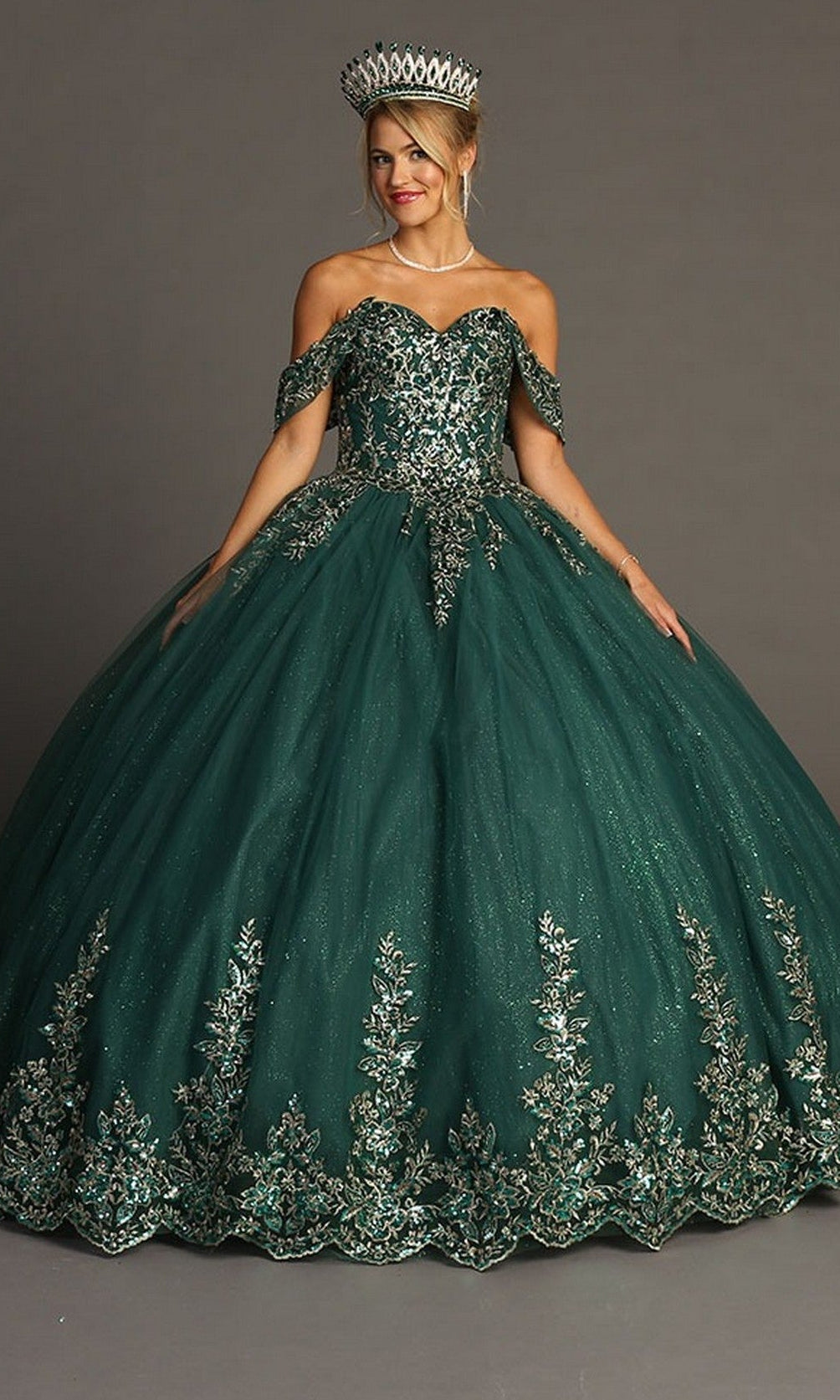A woman wearing the Quinceanera Dress CHF3081 By Chicas in green, an ornate off-the-shoulder ballgown with intricate silver embroidery and a large silver tiara, stands against a plain gray background.