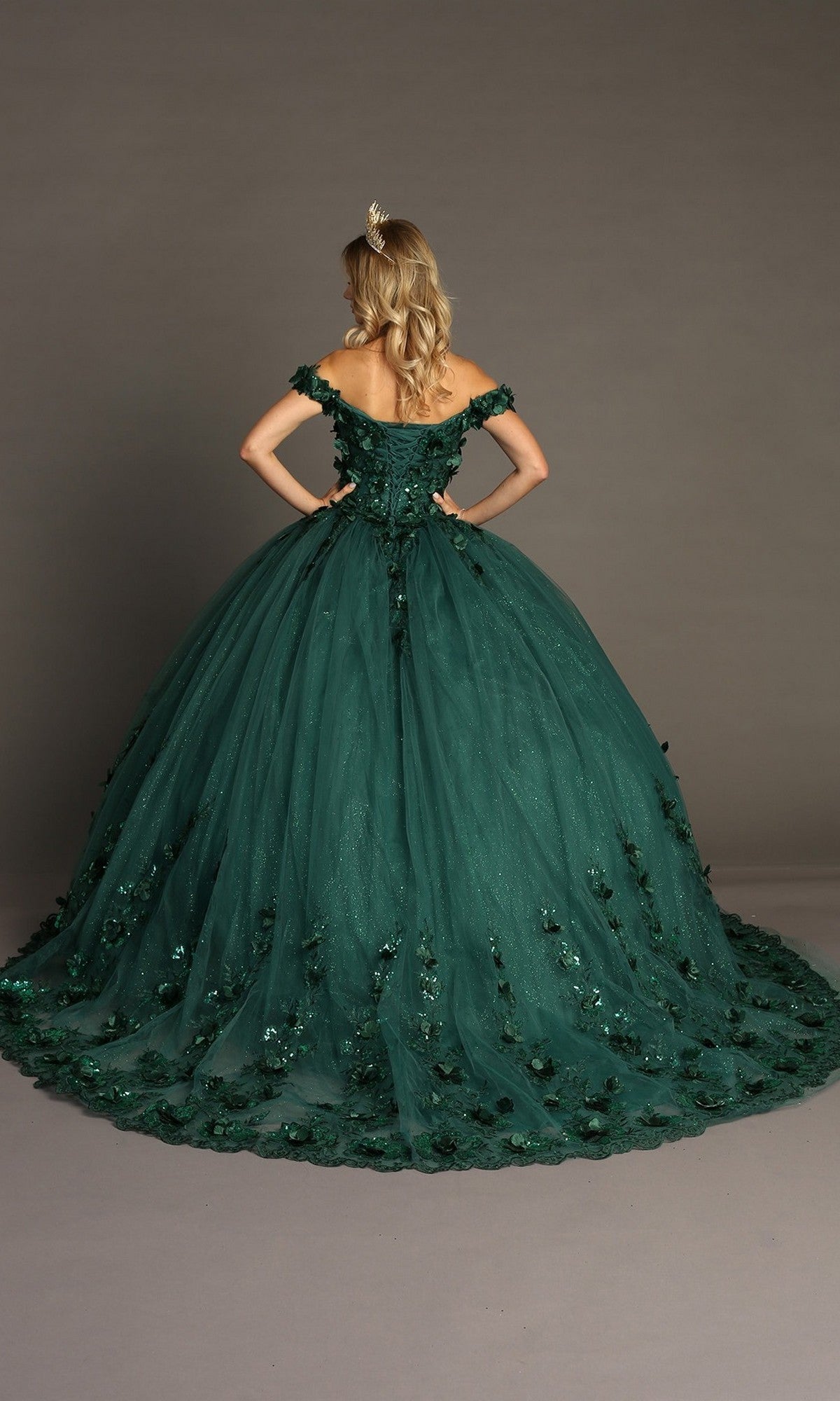 A woman wearing the Quinceanera Dress CHF3082 By Chicas, an off-shoulder dark green gown featuring a long train and intricate 3D floral appliques, stands with hands on hips, facing away.