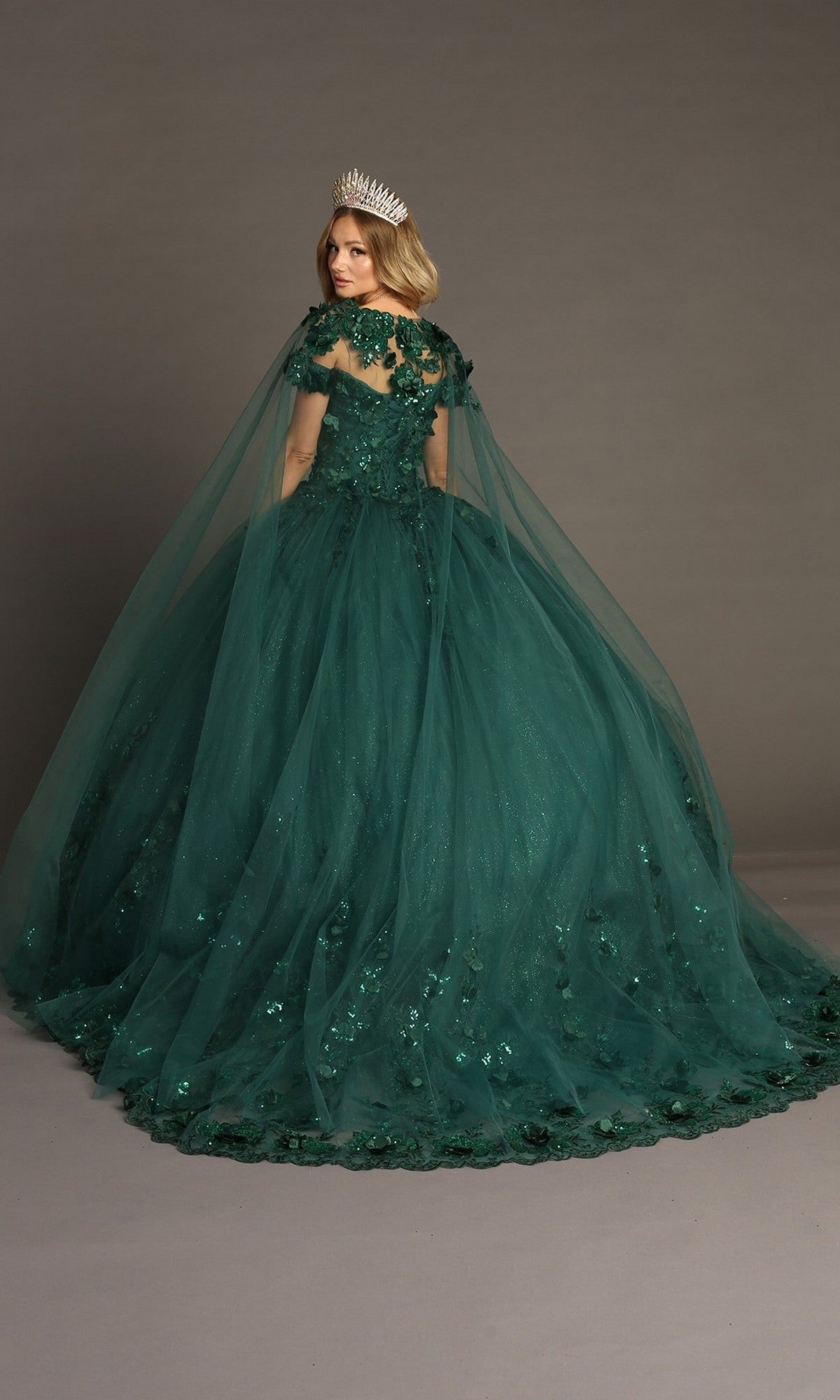 A woman wearing the Quinceanera Dress CHF3082 By Chicas—a dark green gown adorned with sequin details, a sheer cape, and a silver crown—stands on a plain backdrop, facing slightly away from the camera. The dress features delicate 3D floral appliques that add an elegant touch to her ensemble.