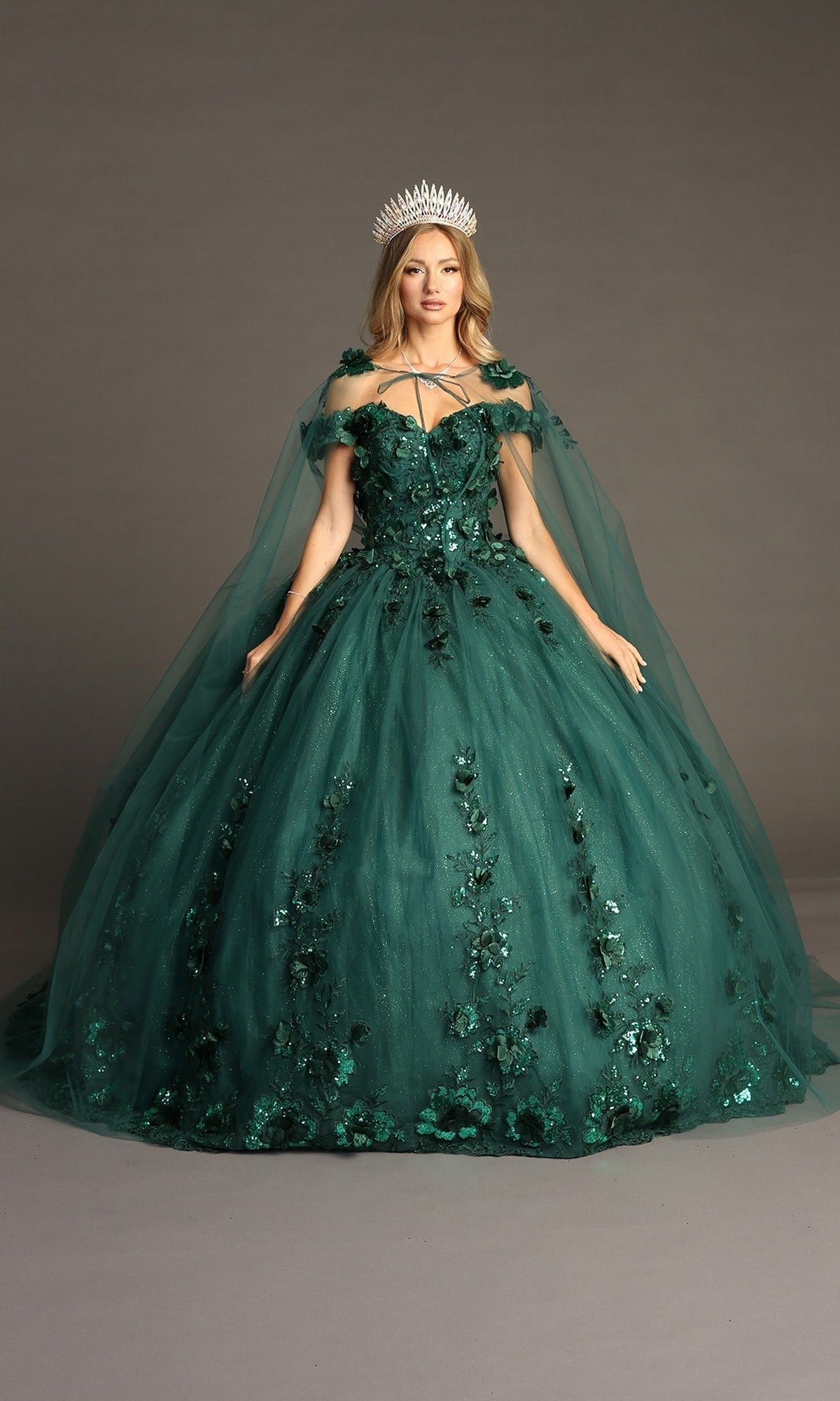 A woman wears the Quinceanera Dress CHF3082 By Chicas in green, adorned with 3D floral appliques and featuring a sheer cape and a silver crown, as she stands against a plain gray background.